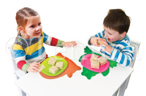 Two children sitting at a white table eating sandwiches, each using colorful animal-shaped Tenura non-slip mats, an orange cat and a green frog as placemats to keep their plates steady.